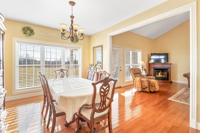 dining space featuring a chandelier, baseboards, vaulted ceiling, light wood finished floors, and a glass covered fireplace