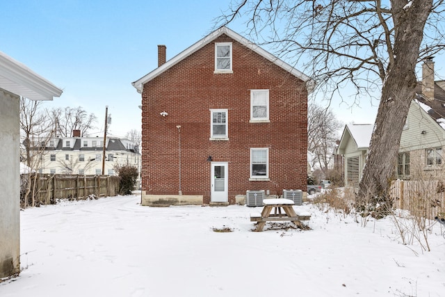 snow covered house with a chimney, fence, cooling unit, and brick siding