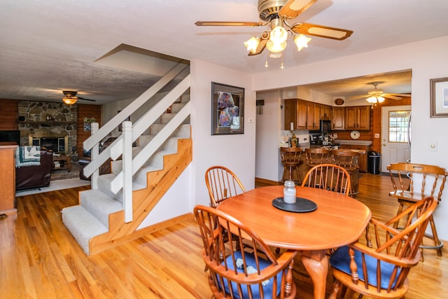 dining area featuring light wood-style floors, ceiling fan, a stone fireplace, and stairs