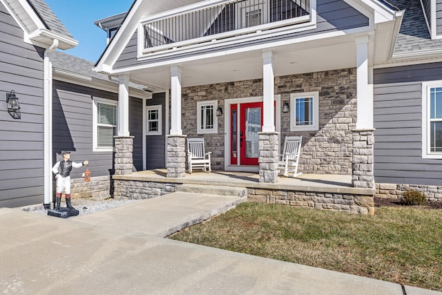 entrance to property with a balcony, stone siding, a porch, and roof with shingles