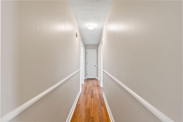 corridor featuring light wood-style floors, baseboards, a textured ceiling, and an upstairs landing