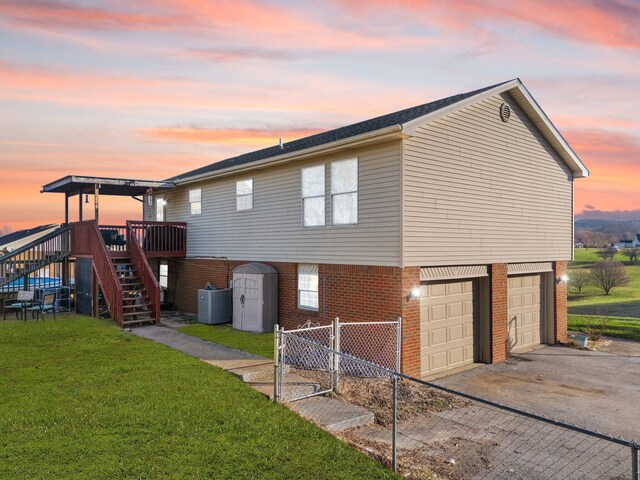 property exterior at dusk with driveway, a yard, brick siding, central AC unit, and stairs