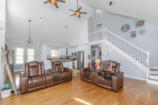 living area featuring baseboards, light wood-style flooring, stairway, high vaulted ceiling, and ceiling fan with notable chandelier