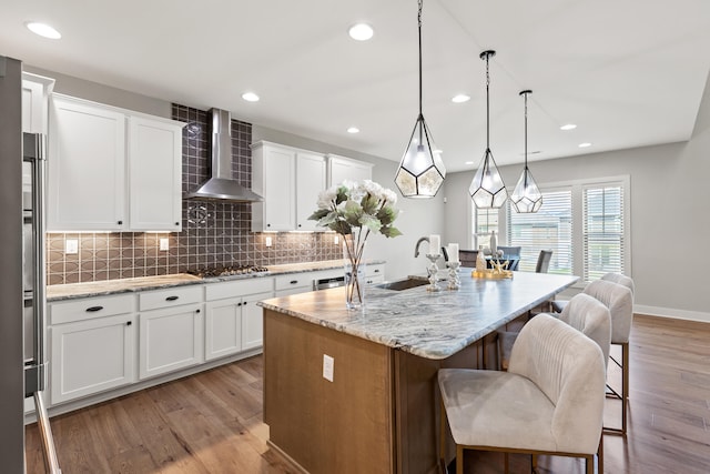 kitchen featuring white cabinets, light stone counters, decorative light fixtures, a kitchen island with sink, and wall chimney range hood