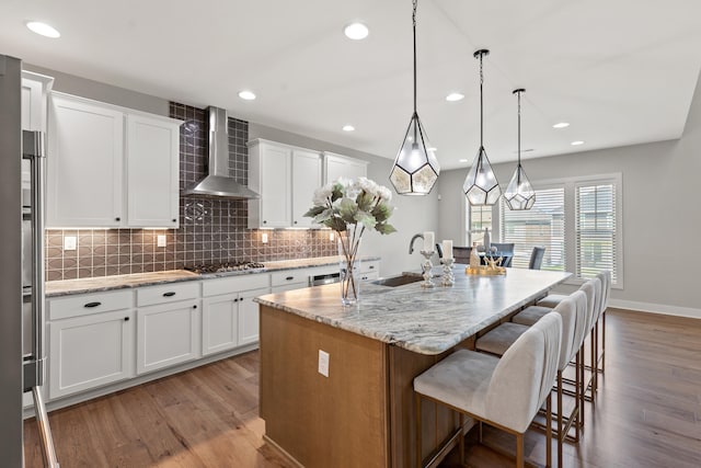 kitchen with white cabinets, a center island with sink, light stone countertops, wall chimney exhaust hood, and decorative light fixtures