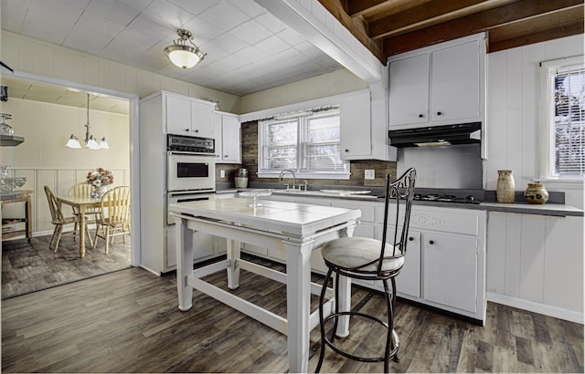 kitchen featuring white cabinets, white double oven, under cabinet range hood, dark wood-type flooring, and a decorative wall