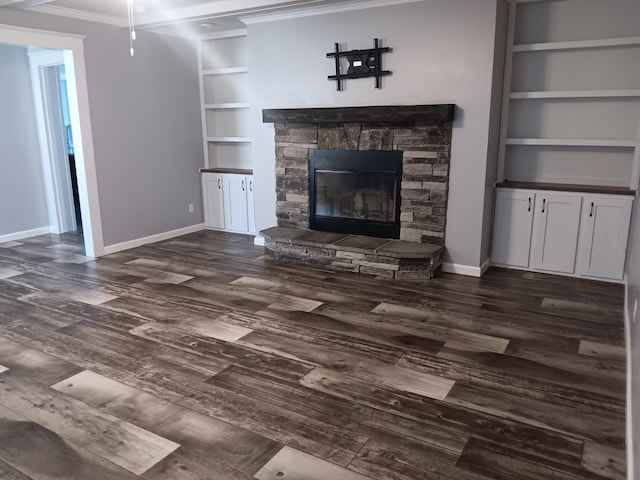 unfurnished living room with a stone fireplace, built in shelves, dark wood-type flooring, baseboards, and ornamental molding