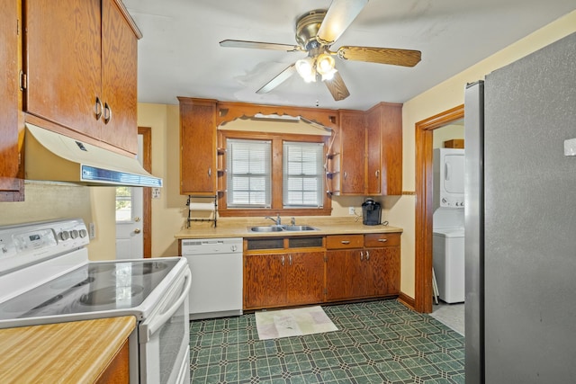 kitchen featuring white appliances, brown cabinetry, under cabinet range hood, stacked washing maching and dryer, and a sink