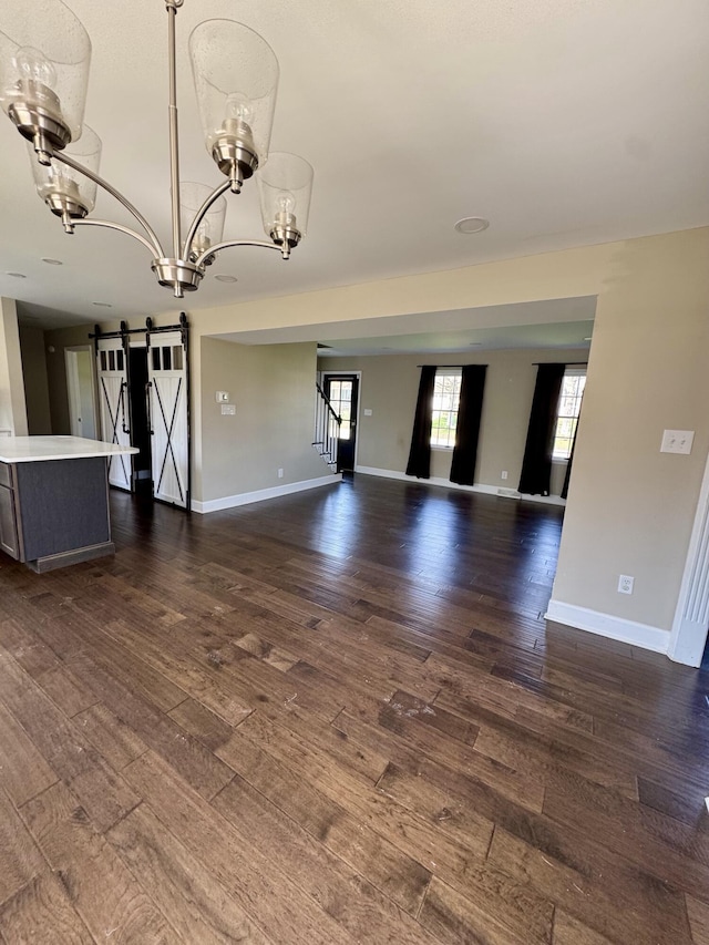 unfurnished living room featuring baseboards, a notable chandelier, a barn door, and dark wood-style floors