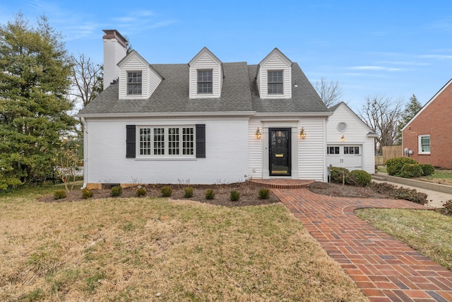 new england style home featuring brick siding, a chimney, roof with shingles, an attached garage, and a front yard