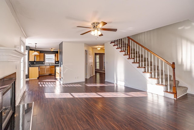 unfurnished living room featuring dark wood-style floors, ceiling fan, stairway, and a glass covered fireplace