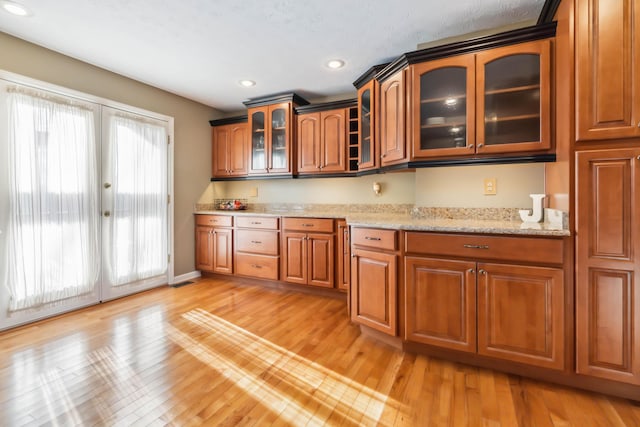 kitchen with recessed lighting, brown cabinets, light stone countertops, light wood finished floors, and glass insert cabinets