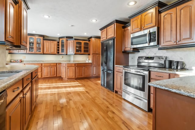 kitchen featuring stainless steel appliances, light wood finished floors, brown cabinetry, and glass insert cabinets