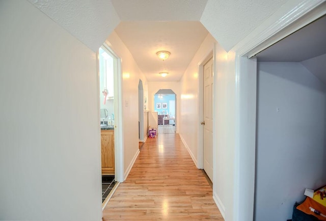 hallway featuring arched walkways, a textured ceiling, baseboards, and light wood-style floors