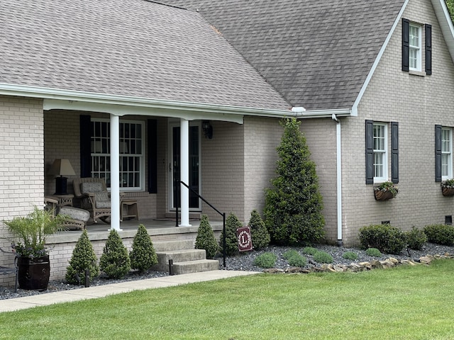 view of exterior entry featuring a shingled roof, a yard, a porch, and brick siding