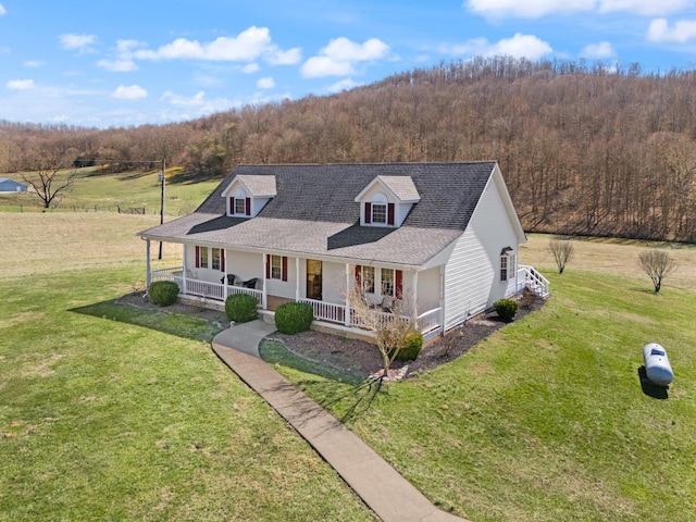 farmhouse with a front lawn, a porch, a view of trees, and roof with shingles