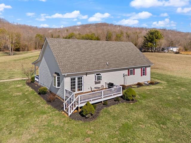 rear view of house with a deck, roof with shingles, a lawn, and a wooded view