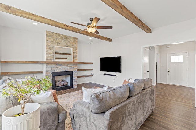 living area with beam ceiling, ceiling fan, a stone fireplace, wood finished floors, and baseboards