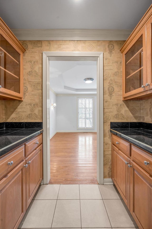 kitchen featuring light tile patterned floors, ornamental molding, dark countertops, glass insert cabinets, and wallpapered walls