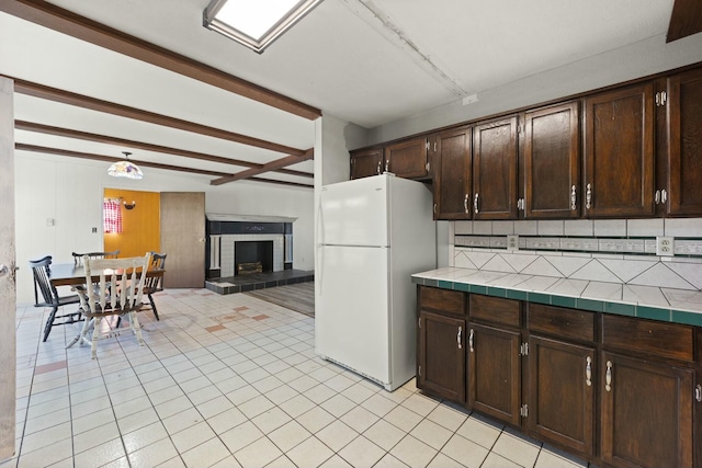 kitchen with dark brown cabinetry, light tile patterned floors, a fireplace with raised hearth, tile countertops, and freestanding refrigerator