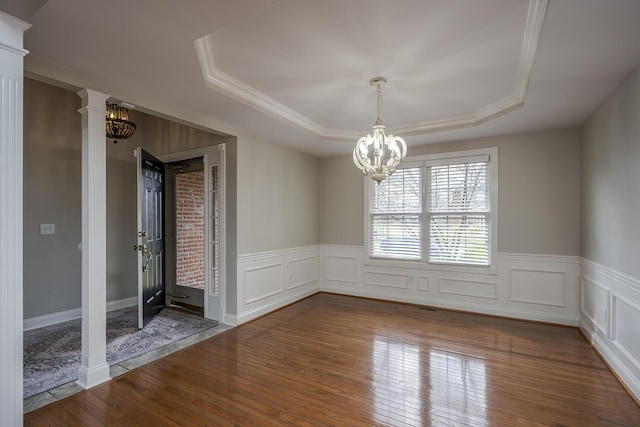 unfurnished room featuring a tray ceiling, wood-type flooring, wainscoting, a chandelier, and ornate columns