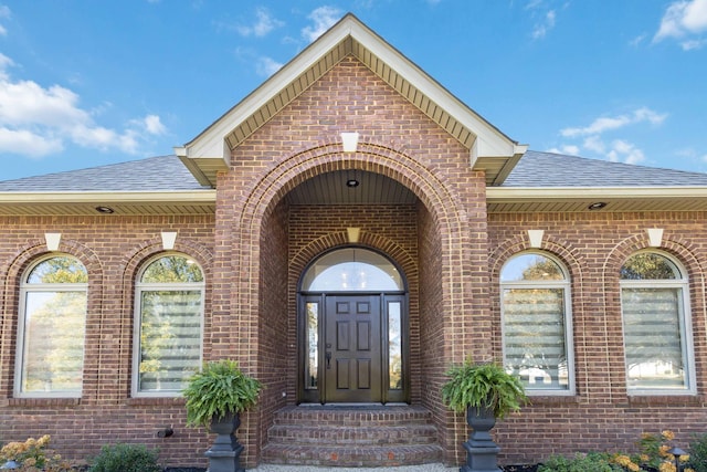 view of exterior entry with a shingled roof and brick siding