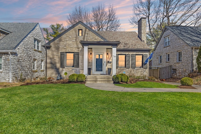 view of front of property featuring stone siding, roof with shingles, a lawn, and a chimney