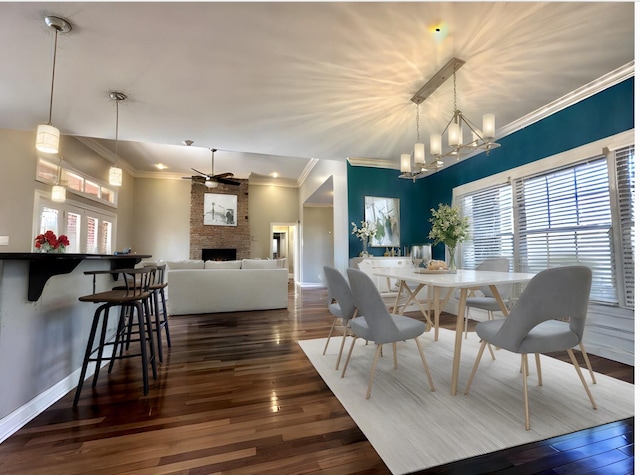 dining area with a large fireplace, ceiling fan with notable chandelier, dark wood-style flooring, baseboards, and ornamental molding