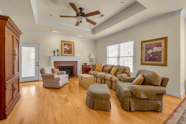 living room with a tray ceiling, a brick fireplace, crown molding, and light wood-style flooring