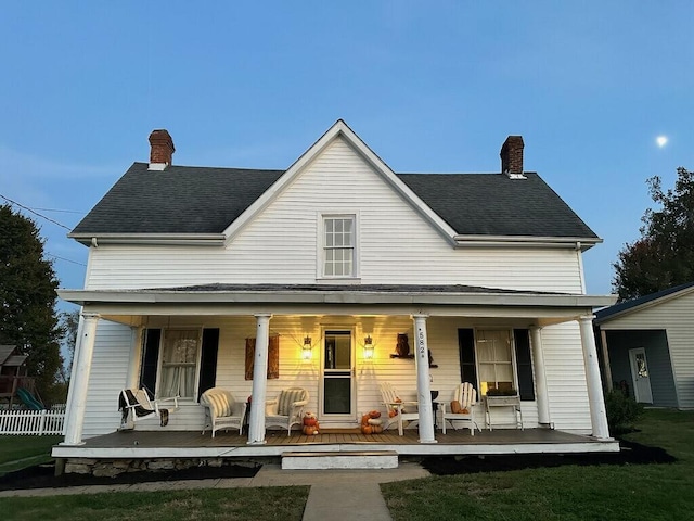 farmhouse-style home with covered porch, a shingled roof, a chimney, and fence