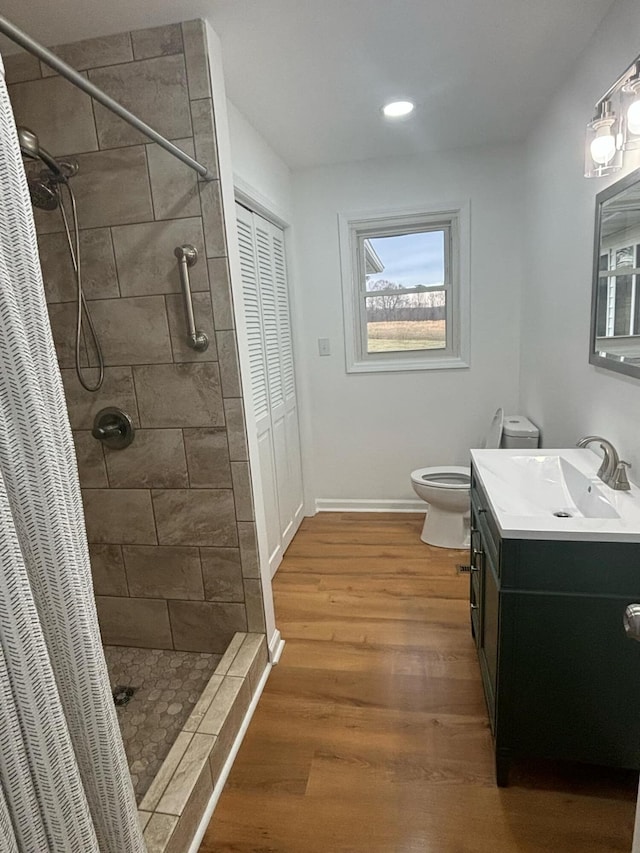 bathroom with toilet, wood finished floors, vanity, baseboards, and tiled shower