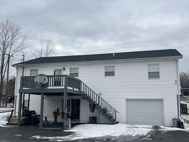 snow covered back of property featuring a garage, a shingled roof, a wooden deck, and stairs