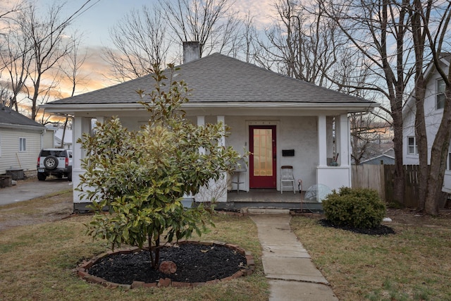 bungalow-style home with a yard, stucco siding, a shingled roof, covered porch, and fence