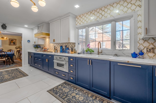 kitchen with light stone counters, appliances with stainless steel finishes, a sink, blue cabinets, and premium range hood