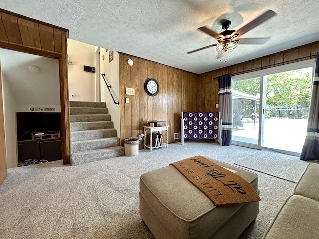 living room featuring a ceiling fan, stairs, carpet flooring, a textured ceiling, and wood walls