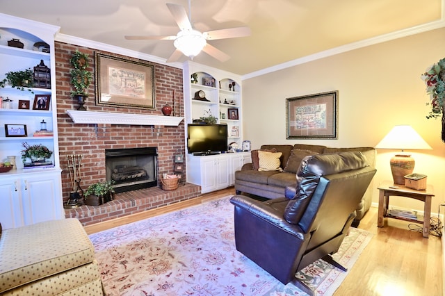 living room with a brick fireplace, a ceiling fan, crown molding, light wood-style floors, and built in shelves