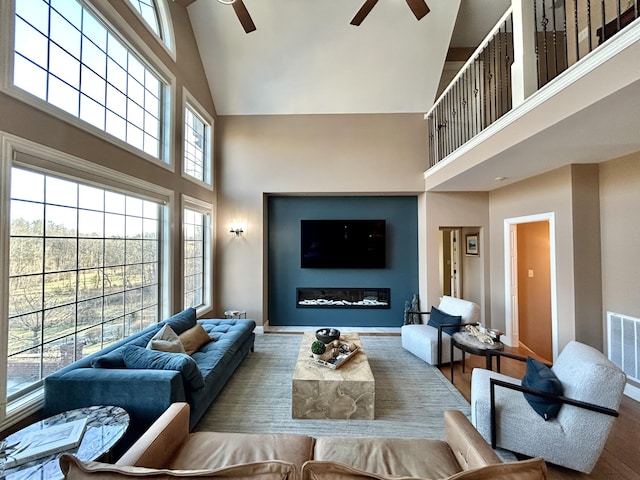 living room with visible vents, a towering ceiling, a ceiling fan, a glass covered fireplace, and wood finished floors