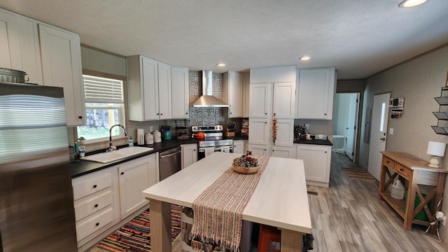 kitchen featuring light wood-style flooring, a sink, appliances with stainless steel finishes, wall chimney exhaust hood, and dark countertops