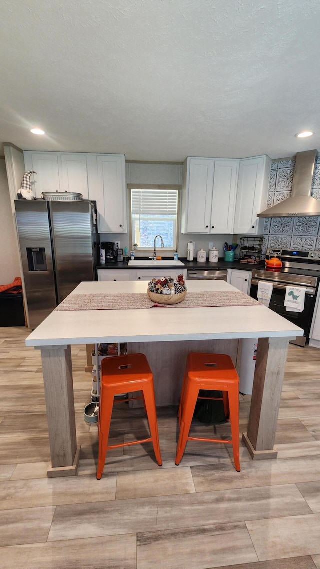 kitchen featuring a center island, appliances with stainless steel finishes, white cabinets, a sink, and wall chimney exhaust hood