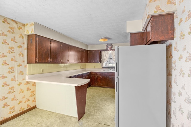 kitchen featuring freestanding refrigerator, a sink, a textured ceiling, a peninsula, and wallpapered walls