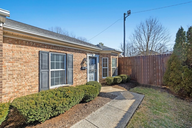 view of exterior entry with roof with shingles, fence, and brick siding