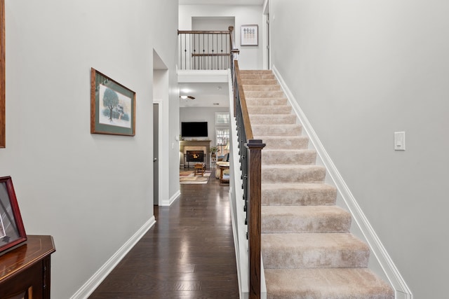 staircase featuring a warm lit fireplace, a high ceiling, baseboards, and wood finished floors