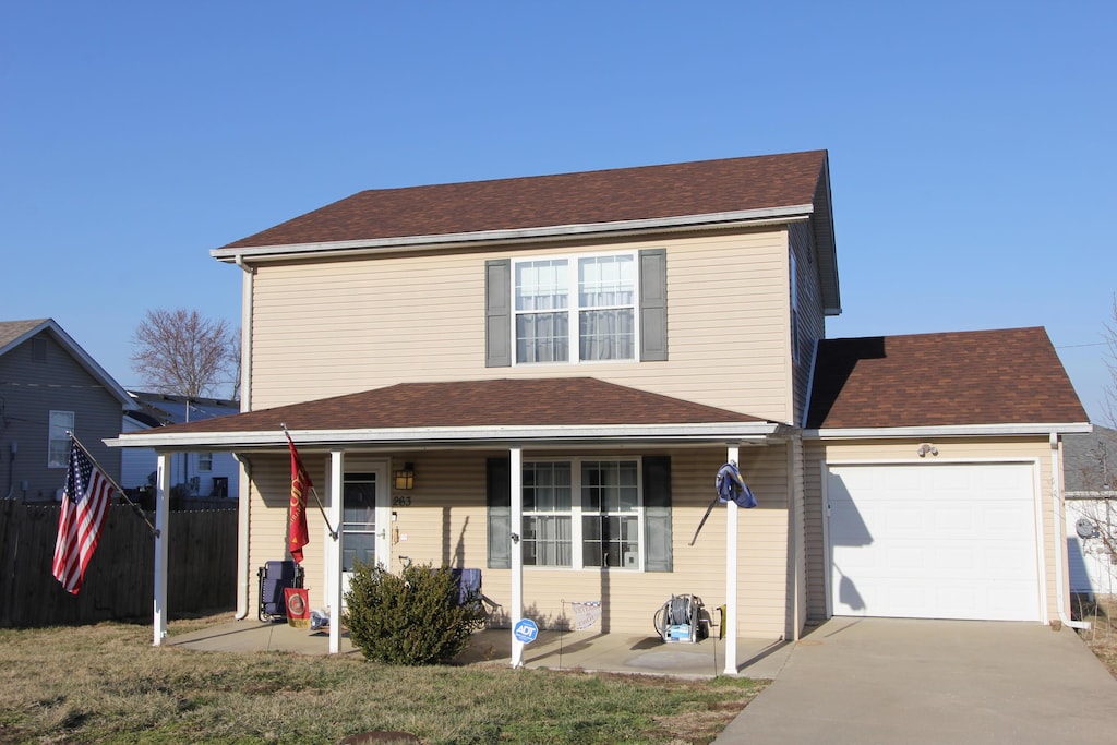 traditional-style home featuring a shingled roof, concrete driveway, an attached garage, covered porch, and fence