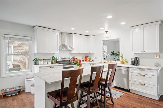 kitchen featuring stainless steel appliances, a breakfast bar, wood finished floors, white cabinetry, and wall chimney exhaust hood