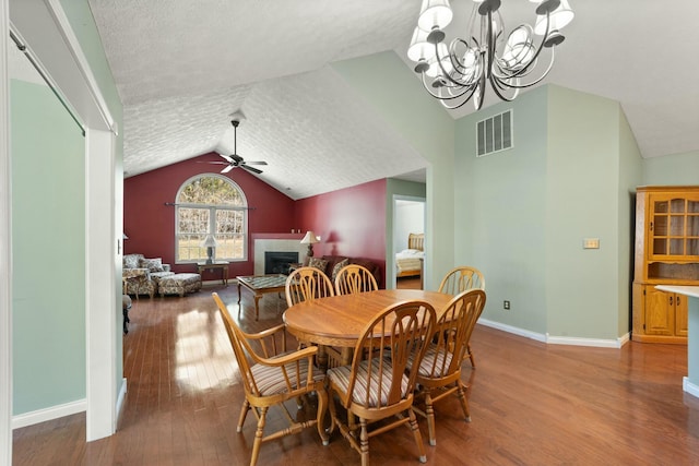 dining space featuring visible vents, wood-type flooring, vaulted ceiling, a fireplace, and ceiling fan with notable chandelier