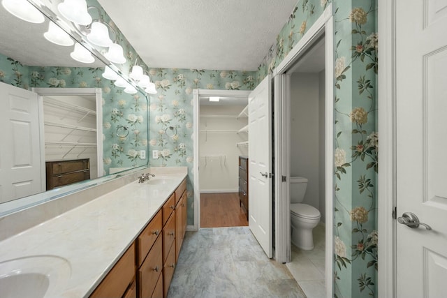 bathroom with double vanity, wallpapered walls, toilet, a textured ceiling, and a sink