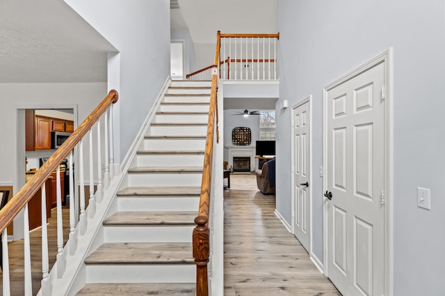 stairs with a ceiling fan, a fireplace, a towering ceiling, and wood finished floors