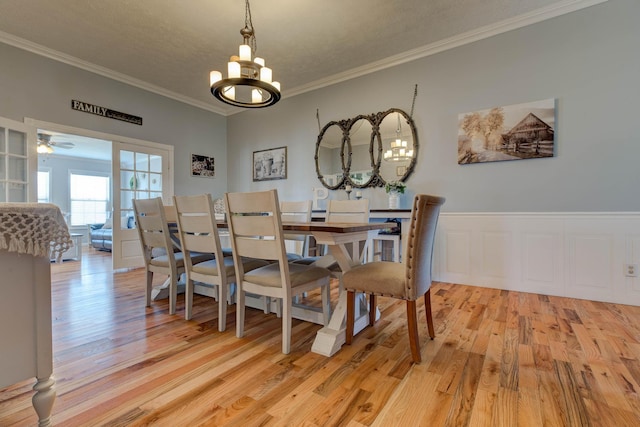 dining space with light wood-style flooring, a decorative wall, a wainscoted wall, ceiling fan with notable chandelier, and crown molding