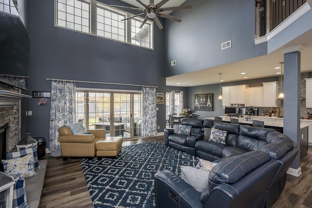 living room featuring baseboards, visible vents, a ceiling fan, dark wood-style flooring, and a stone fireplace