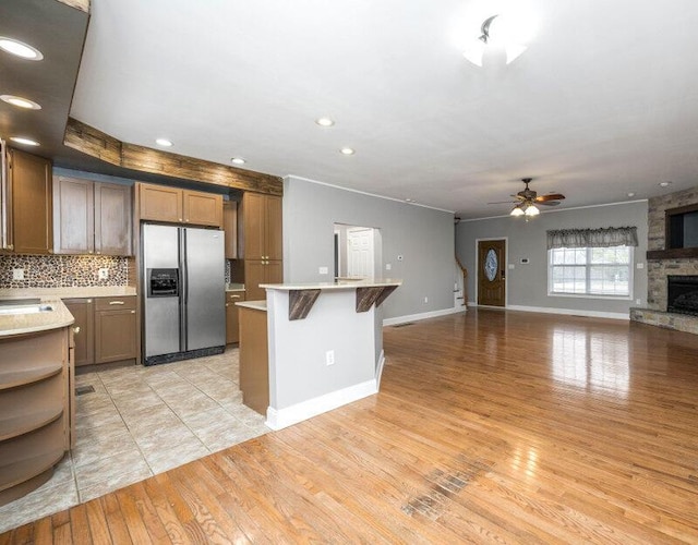 kitchen featuring tasteful backsplash, a ceiling fan, stainless steel fridge with ice dispenser, a stone fireplace, and light wood-style floors